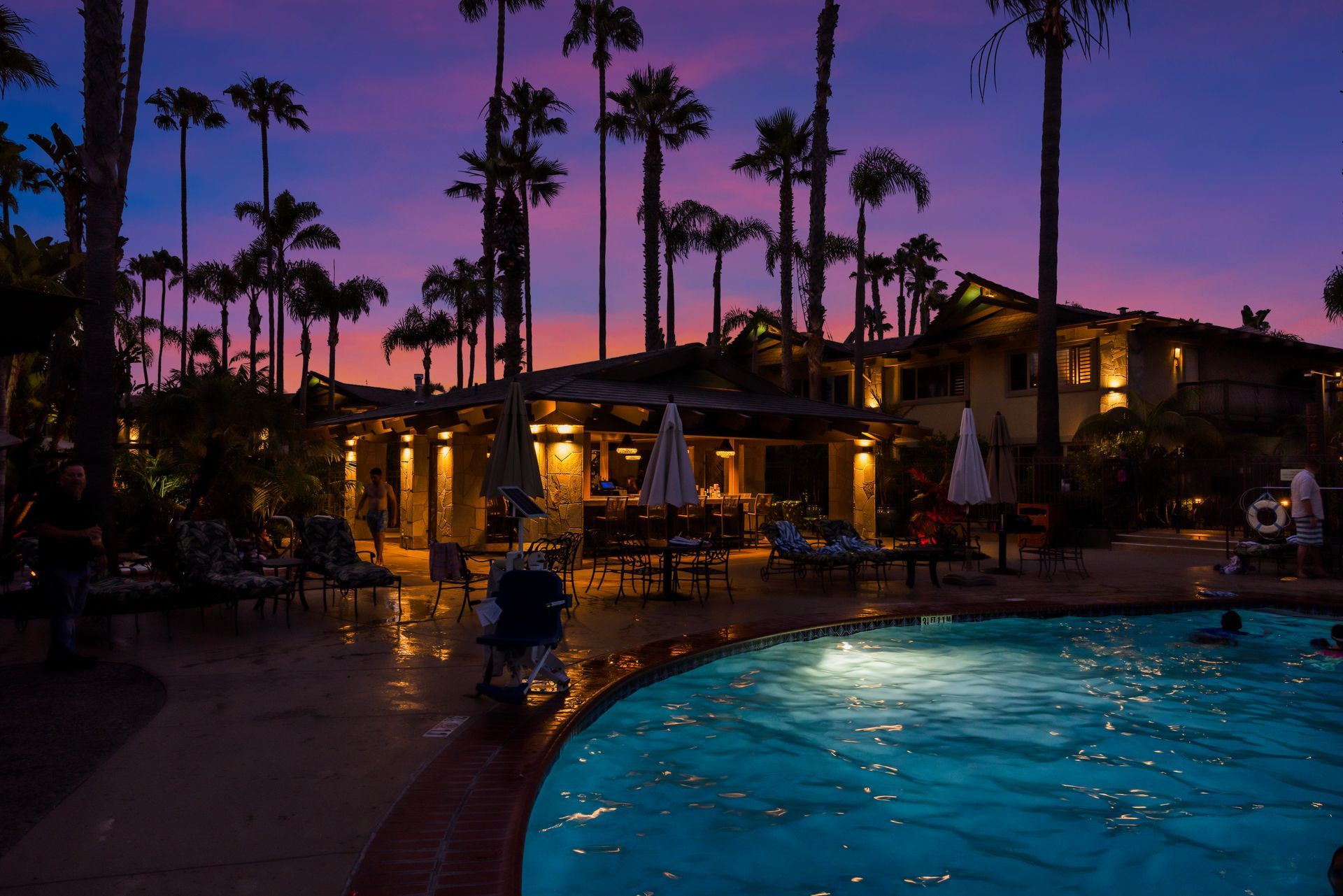 Palm-lined resort pool at dusk with glowing lights and a purple sunset sky