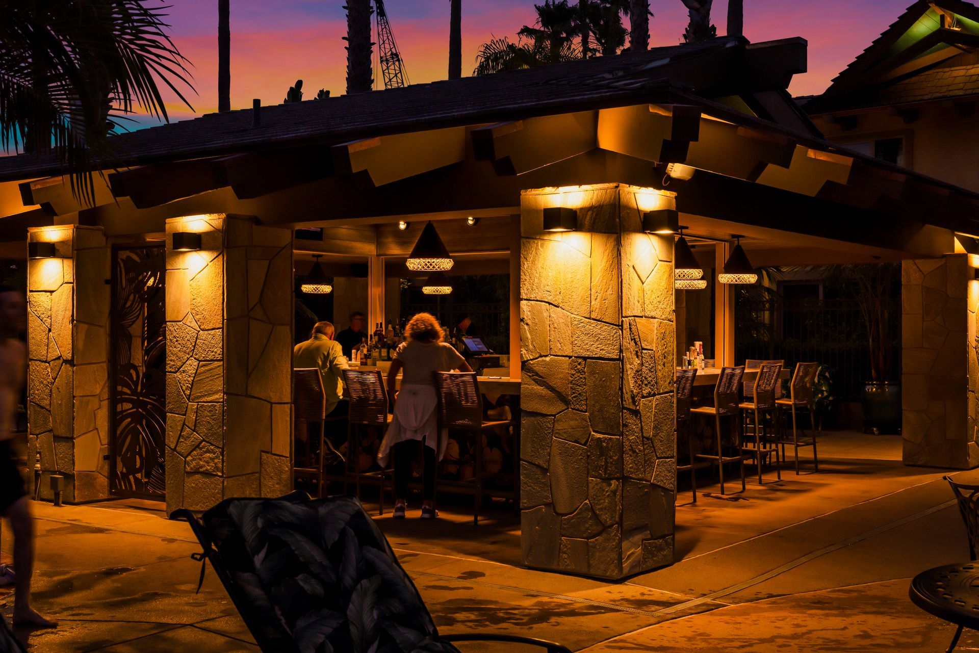Outdoor bar pavilion at dusk with warm lights, stone pillars, and patrons seated inside under a purple sunset sky