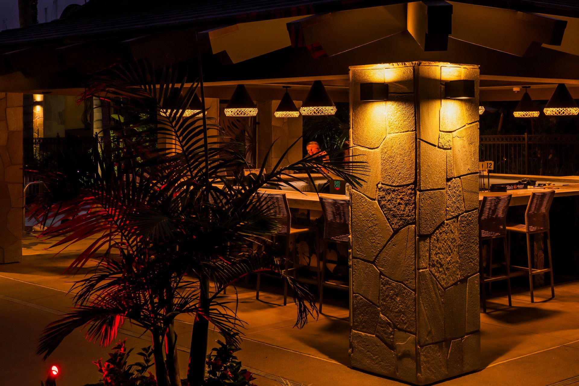 Warmly lit outdoor restaurant patio with stone columns, tables, chairs, and potted plants at night