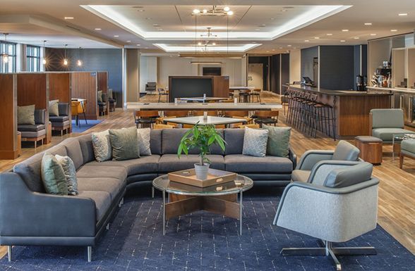 Modern hotel lounge with gray sectional seating, round table, and bar area in the background