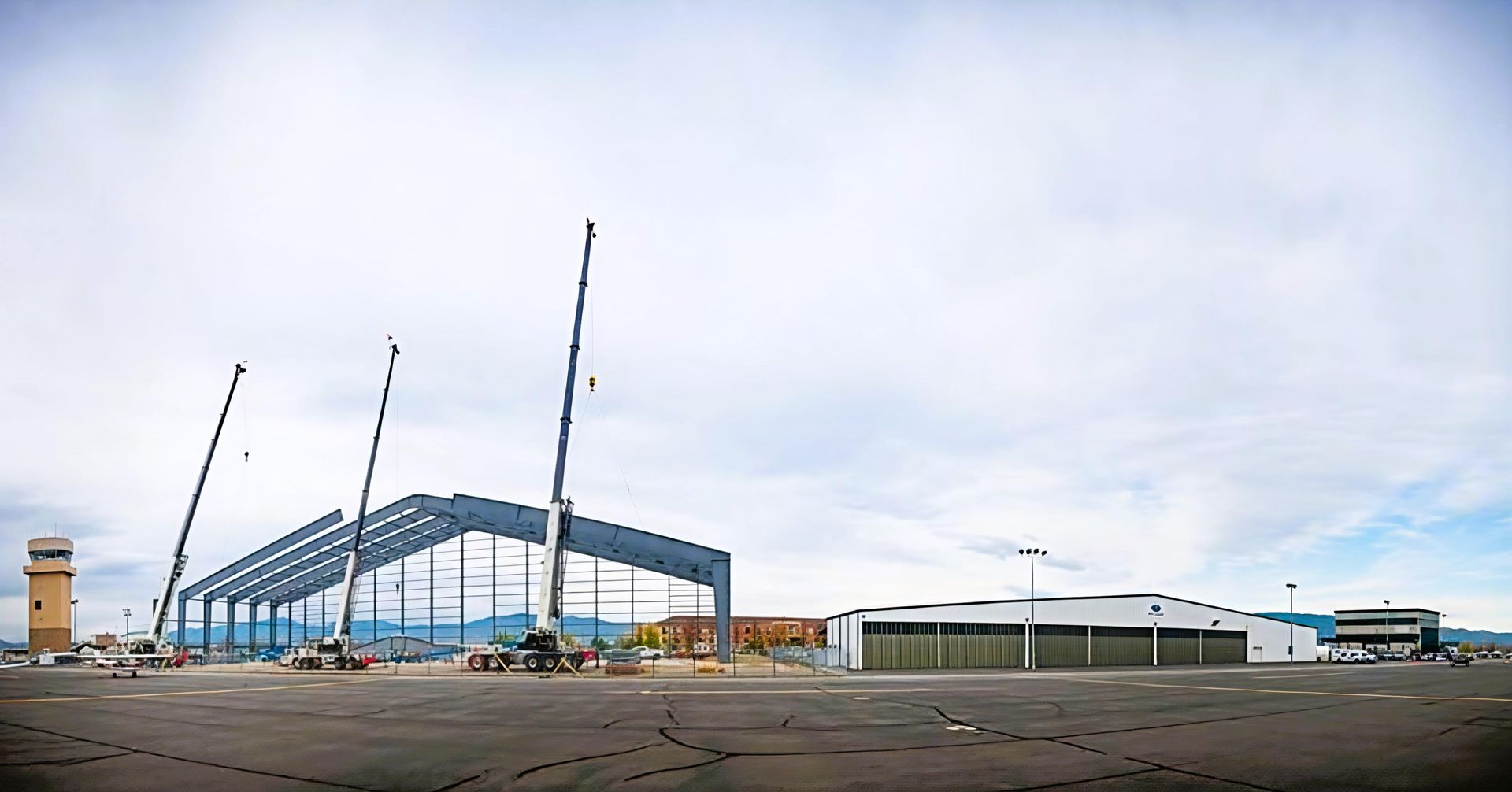 Construction site with steel frame, cranes, and low buildings under a cloudy sky