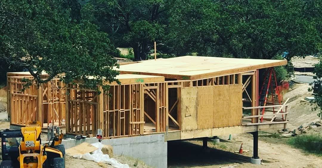 Wood-framed house under construction with a roof, surrounded by trees and a yellow excavator.