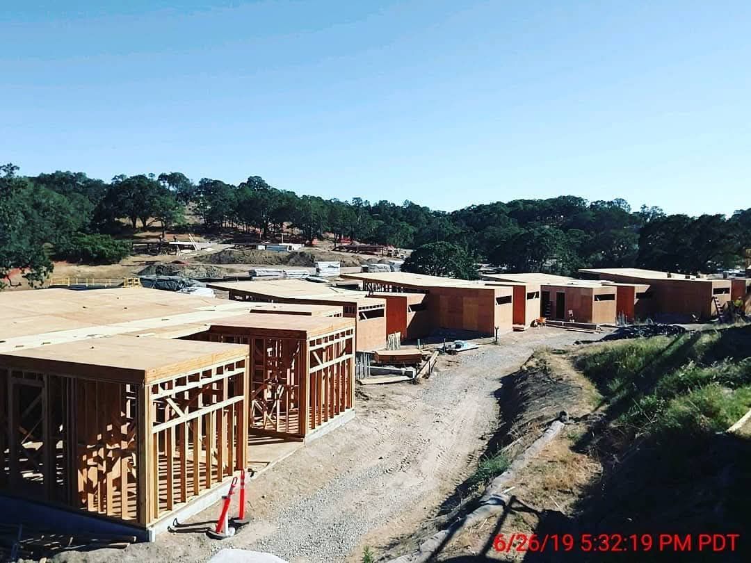 Construction site with wooden framing and concrete foundations along a dirt road under a clear blue sky