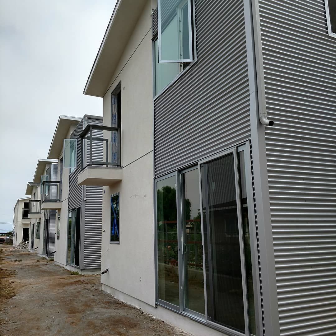 Row of modern gray and white townhouses beside a muddy dirt road, with a cloudy sky.