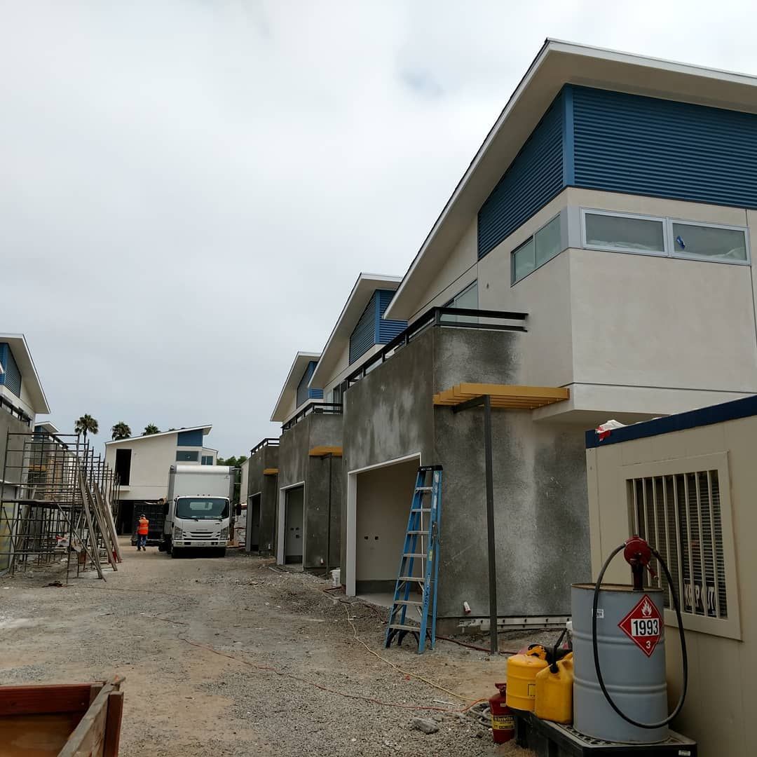 Construction site with row of buildings, gravel road, truck, ladder, and scaffolding under a cloudy sky