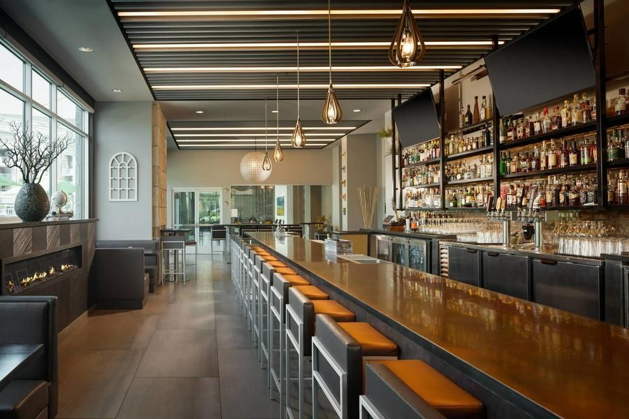 Modern bar interior with a long wooden counter, high stools, and shelves of bottles along the wall
