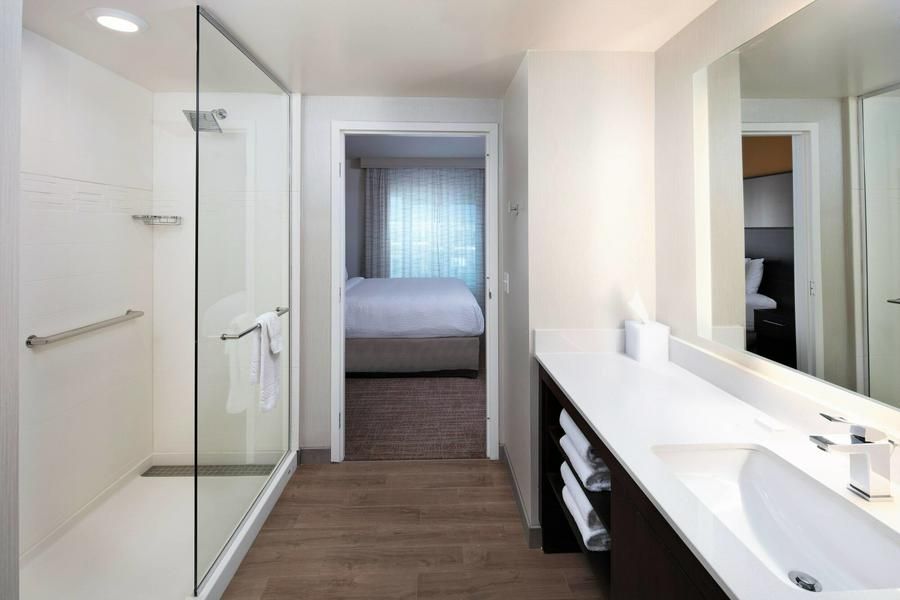 Modern white bathroom with glass shower, vanity, and a hallway view to a bedroom.