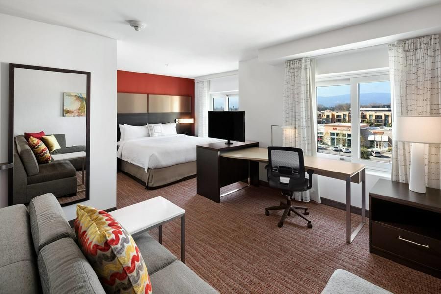 Modern hotel room with bed, desk, armchair, and large window overlooking buildings