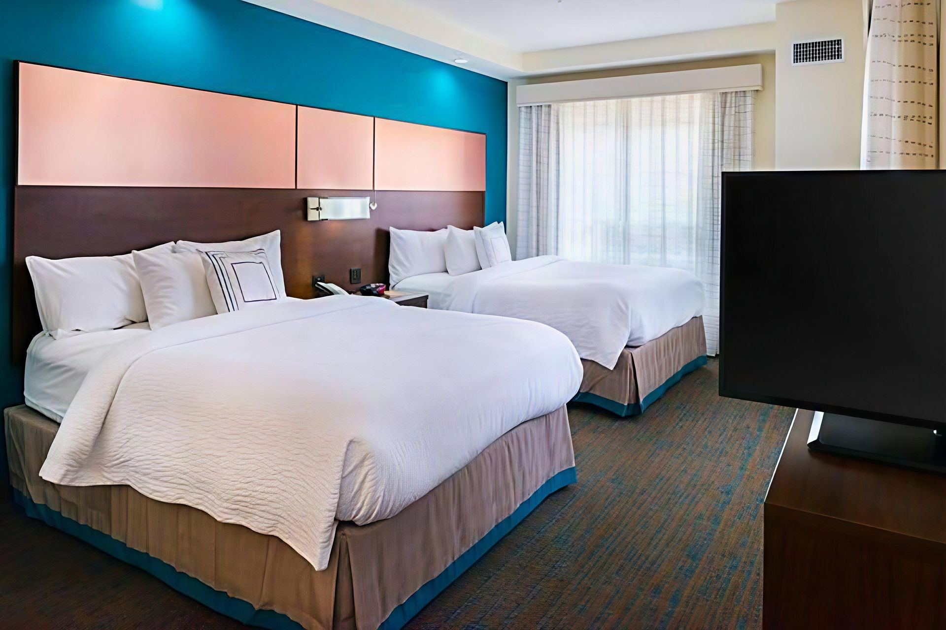 Hotel room with two neatly made queen beds, teal and peach accent wall, TV, and large window with curtains