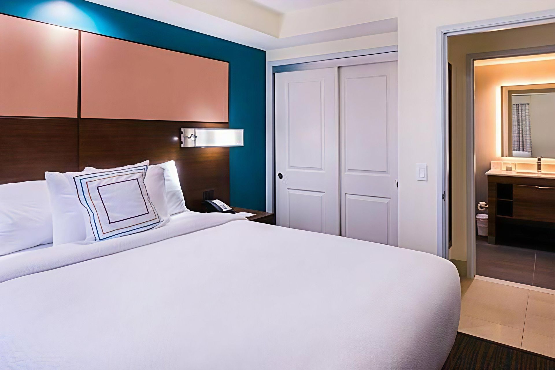 Modern hotel bedroom with a large white bed, pink headboard, and open doorway to a bathroom vanity.