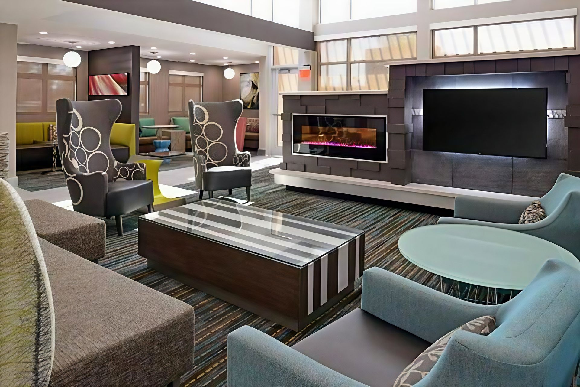 Modern hotel lobby with patterned chairs, striped bench, glass tables, and a fireplace beside a large TV wall