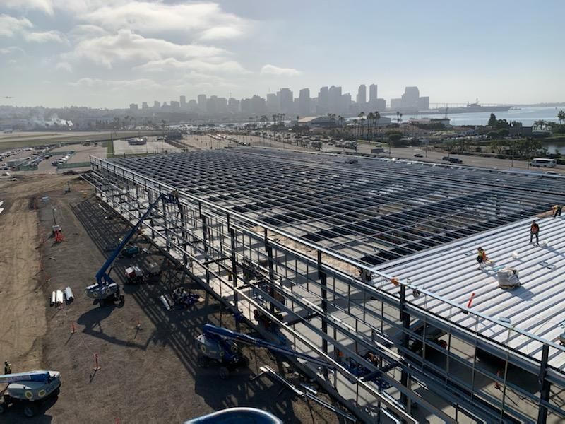 Solar panel canopy under construction in an industrial lot, with city skyline and water in the distance