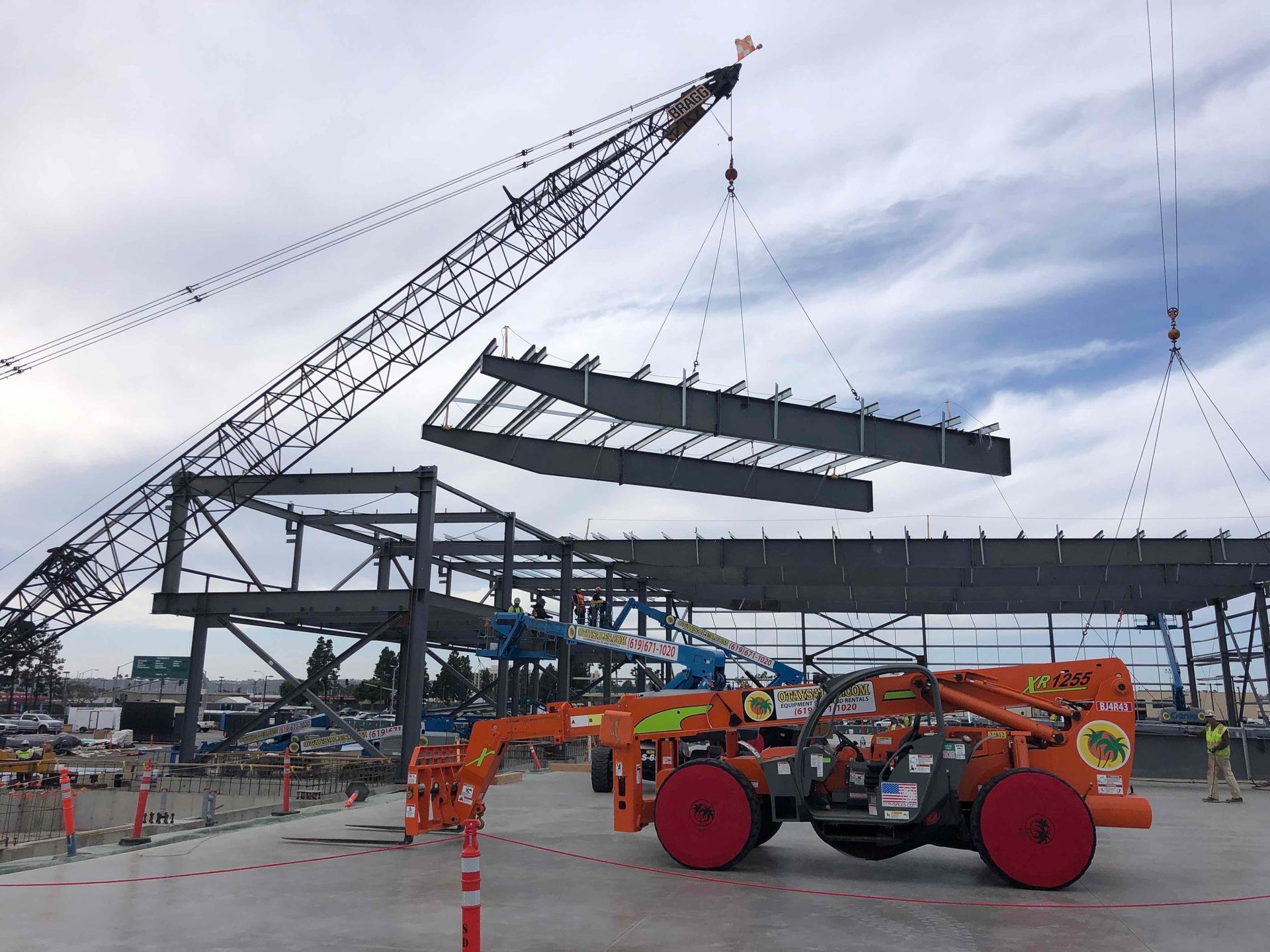 Construction site with a crane lifting steel beams near a partially built structure and orange equipment in front