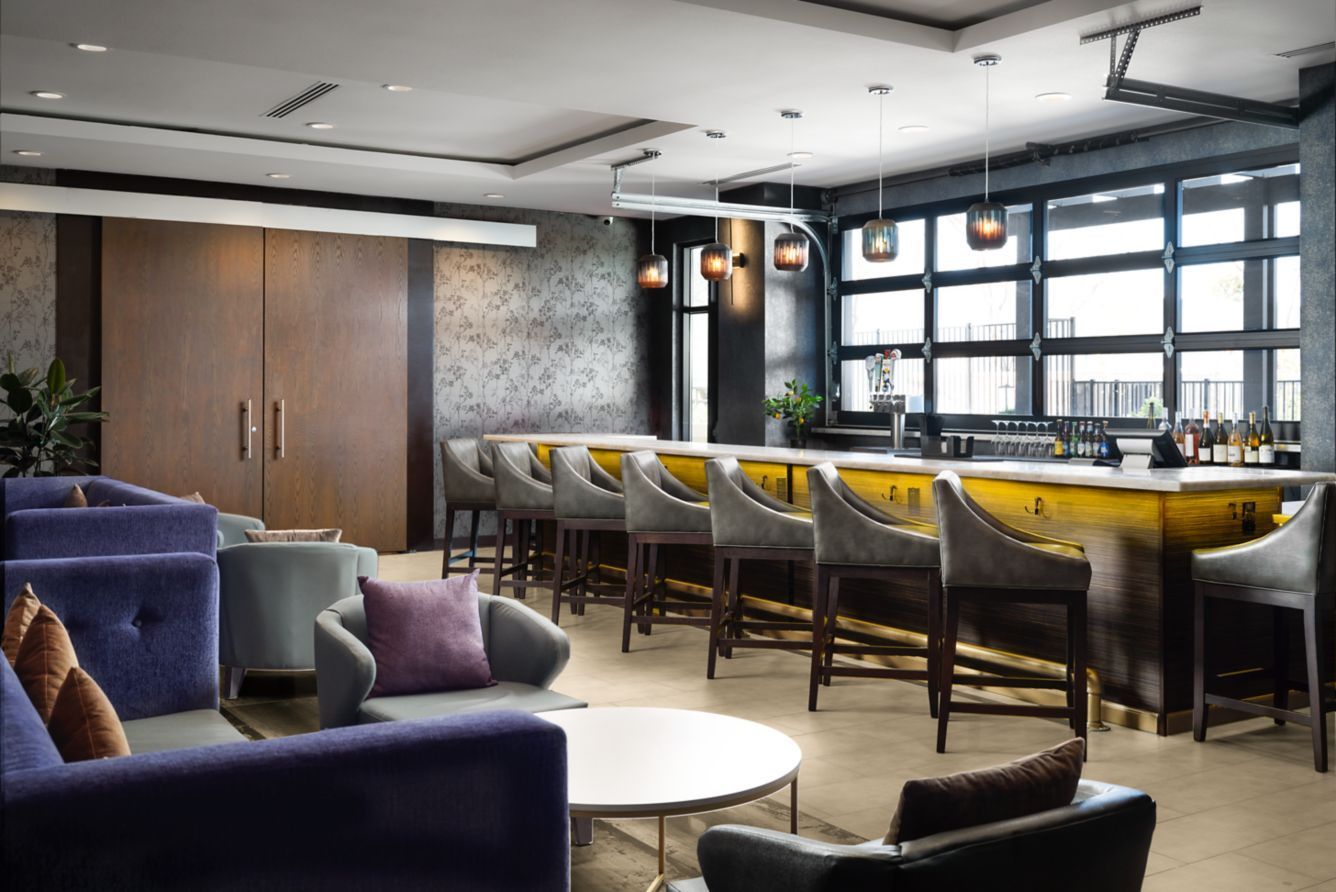 Modern lounge with purple seating, round white tables, and yellow chairs at a long window bar