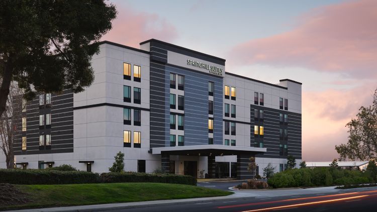 Modern hotel exterior at dusk with lit windows, a covered entrance, and a landscaped driveway.