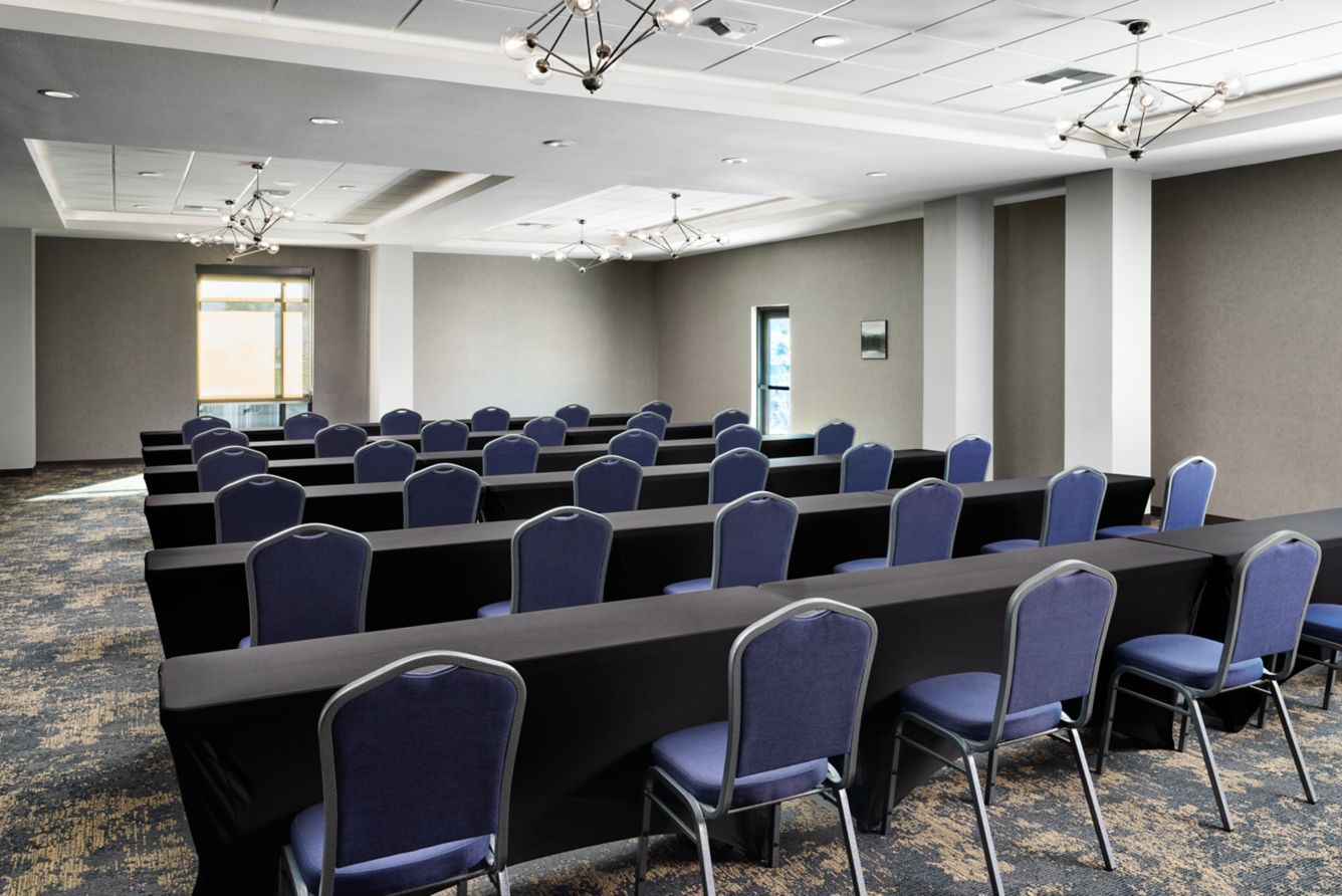 Empty conference room with rows of blue chairs and long tables under ceiling lights.