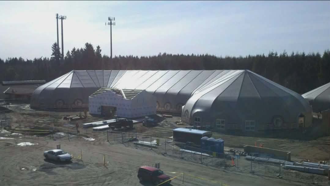 Large silver industrial tent-like structure in a barren field with scattered equipment and trucks under a cloudy sky