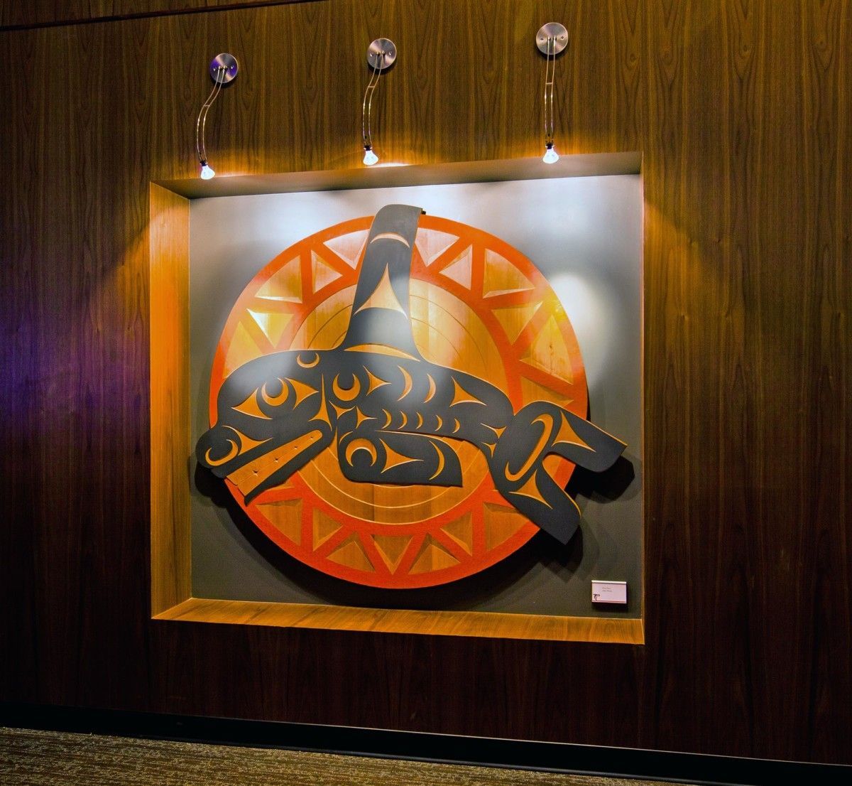 A lit wall-mounted artwork of a stylized black fish on an orange circular background in a wood-paneled room
