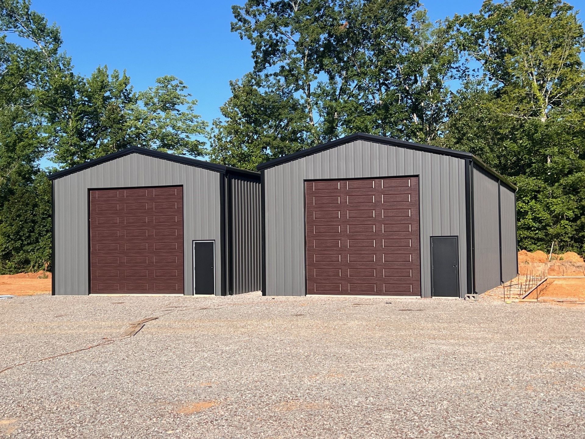 Two gray metal storage sheds with brown doors on a gravel lot, surrounded by trees