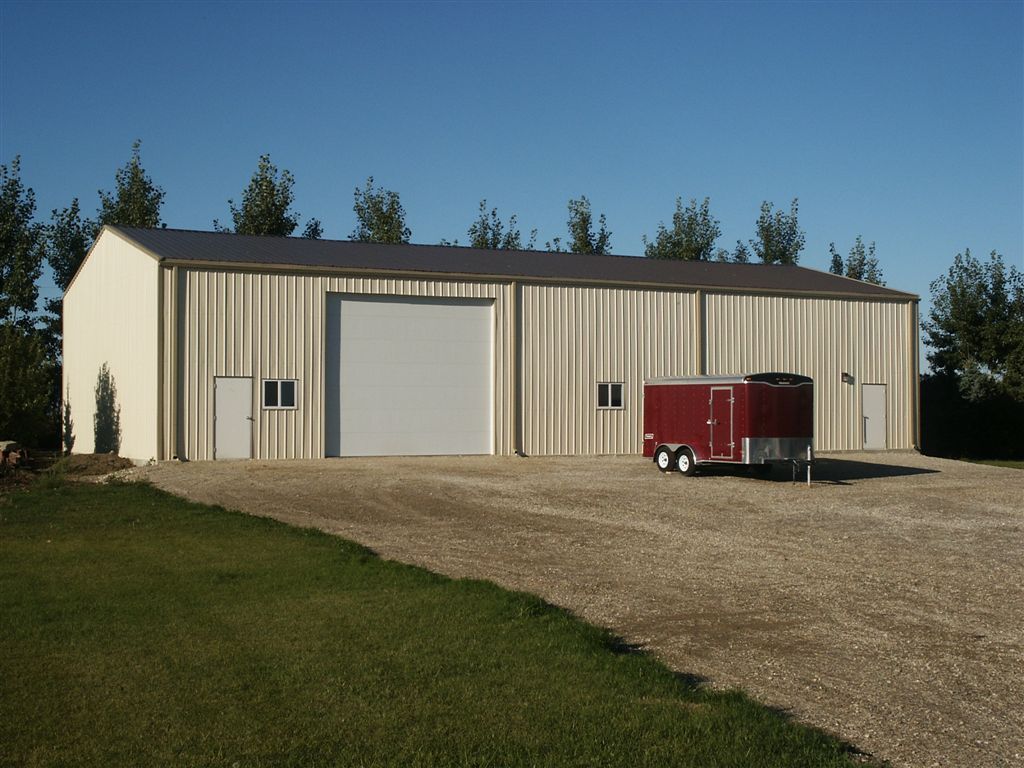 Large beige metal warehouse with a roll-up door and red trailer on a gravel lot under a blue sky