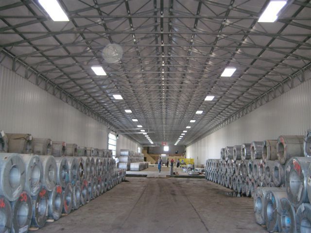 Warehouse interior with rows of stacked metal coils along both sides and a central aisle under high ceiling lights