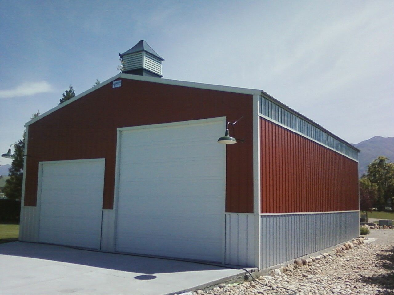 Red and white metal barn with large garage doors on a gravel lot under a blue sky