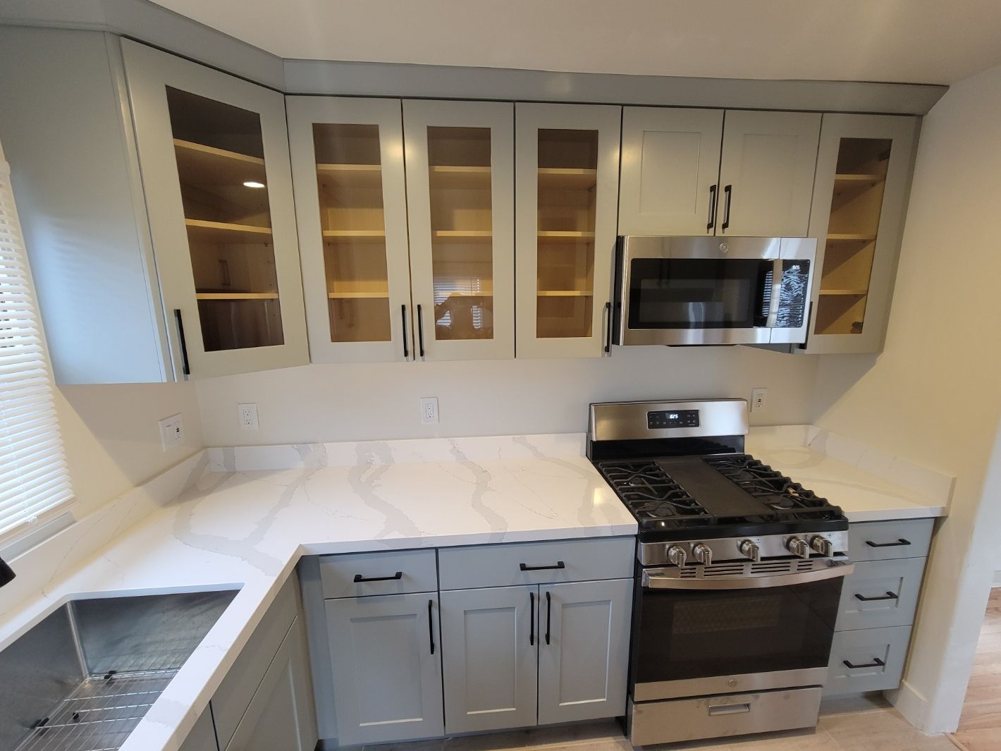 Bright white kitchen with gray cabinets, stainless stove and microwave, and glass-front upper cabinets.