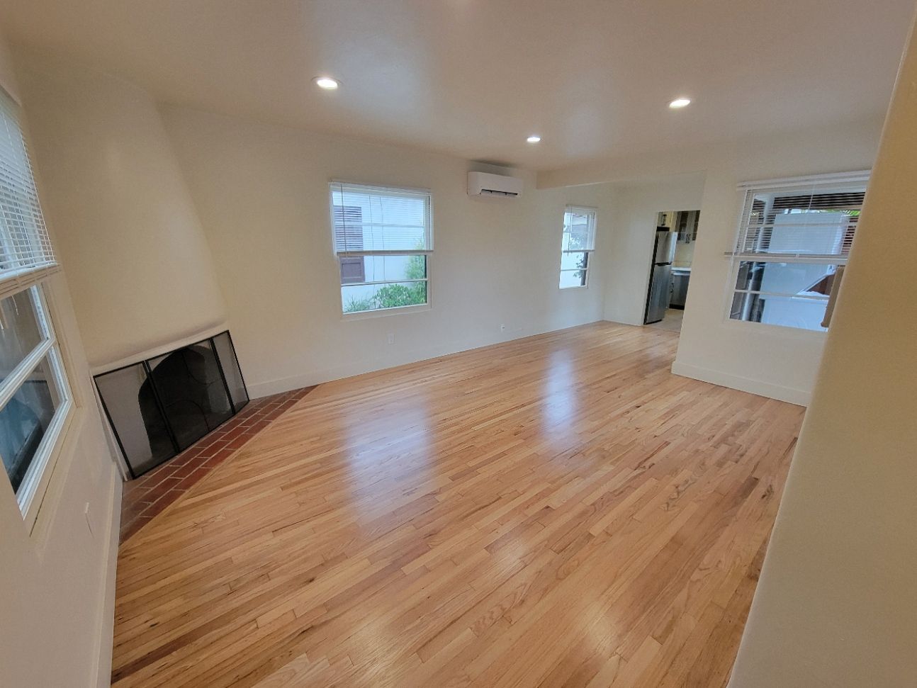 Empty sunlit living room with hardwood floors, white walls, windows, and a fireplace/TV nook