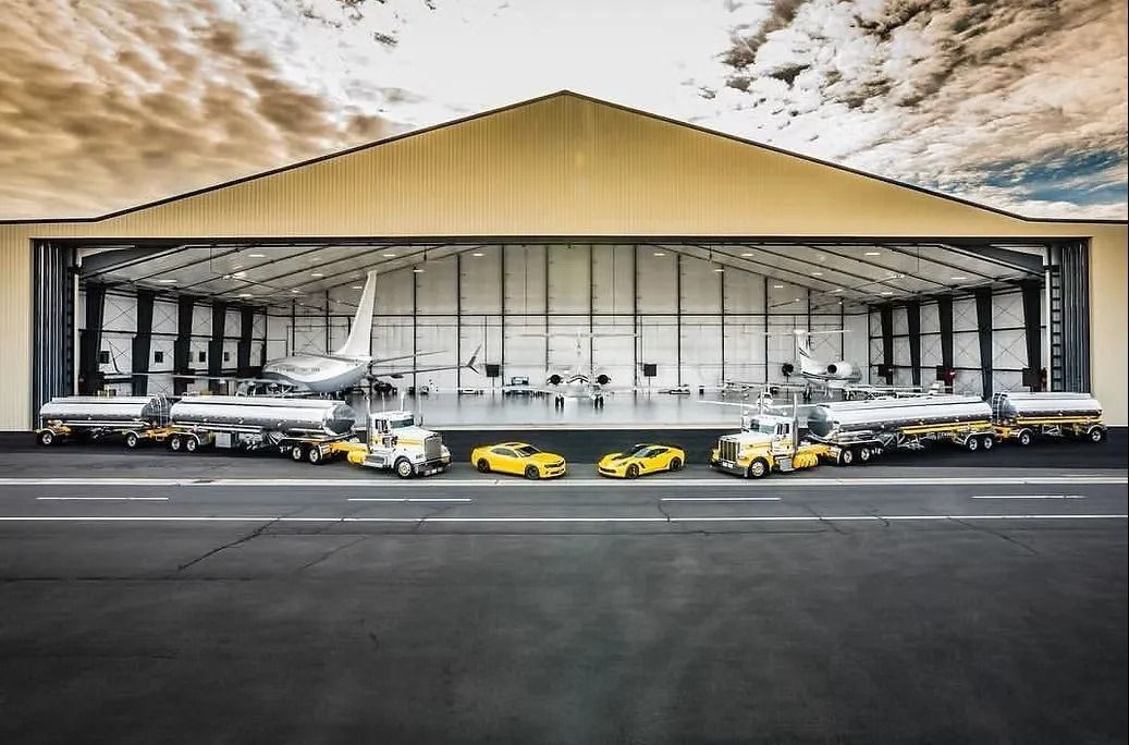 Aircraft hangar with fuel trucks and yellow cars on a tarmac. Planes visible inside the building.