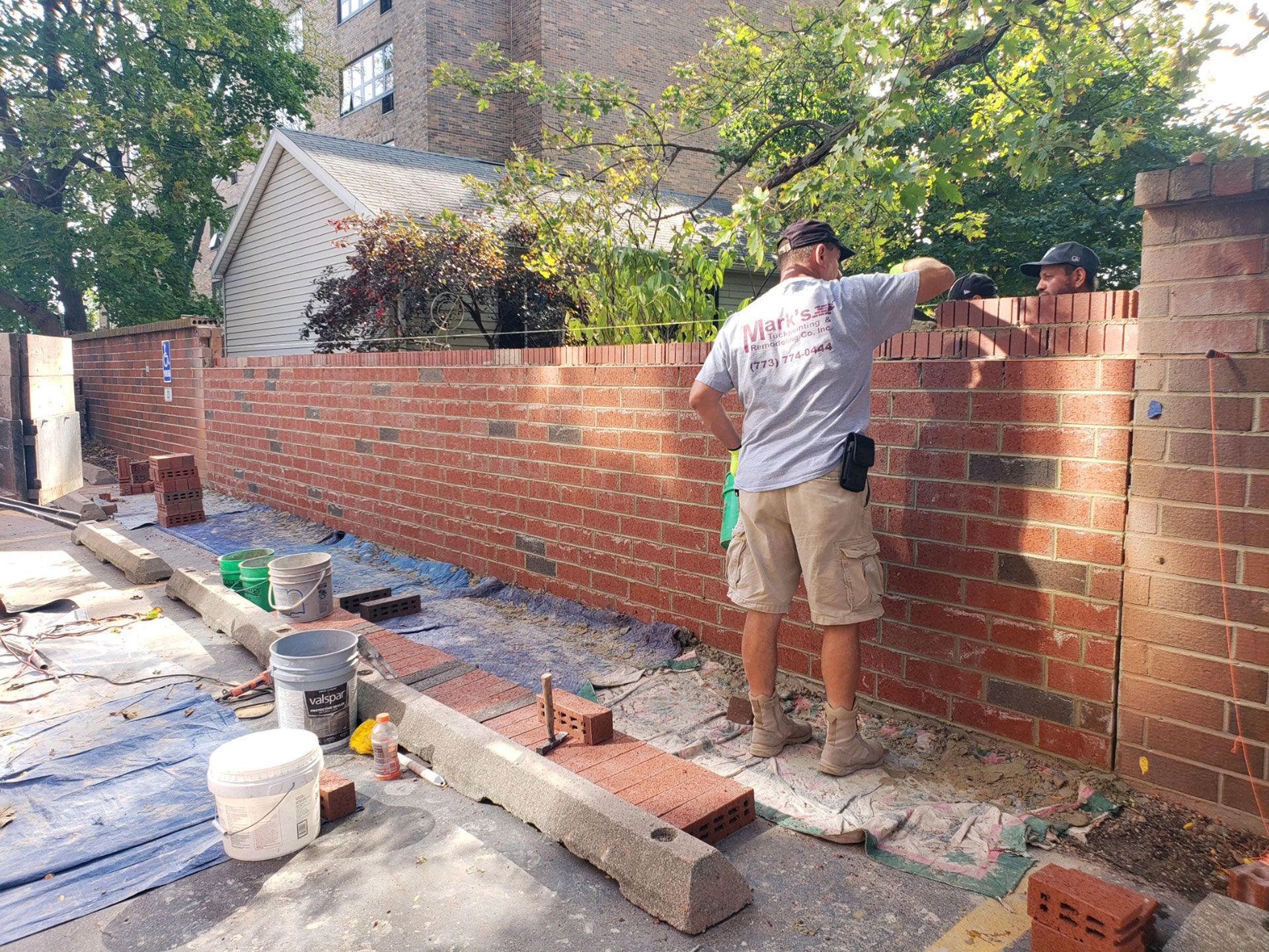 Worker building a red brick wall beside a sidewalk, with tools and materials scattered around.