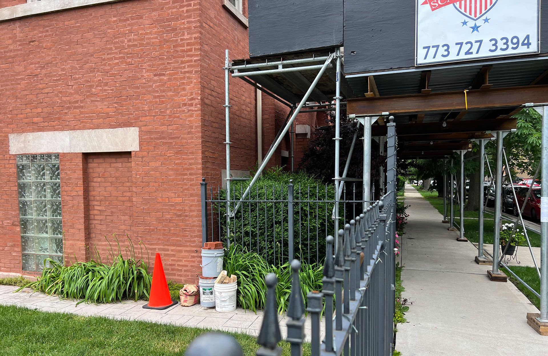 Sidewalk beside brick building with metal scaffolding, black fence, orange cones, and covered walkway