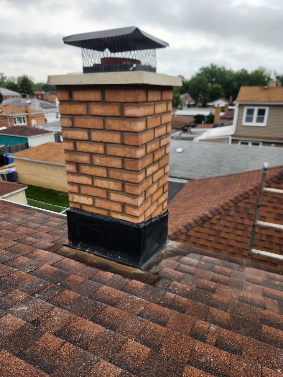 Brick chimney on a shingled rooftop with a metal cap, overlooking nearby houses.