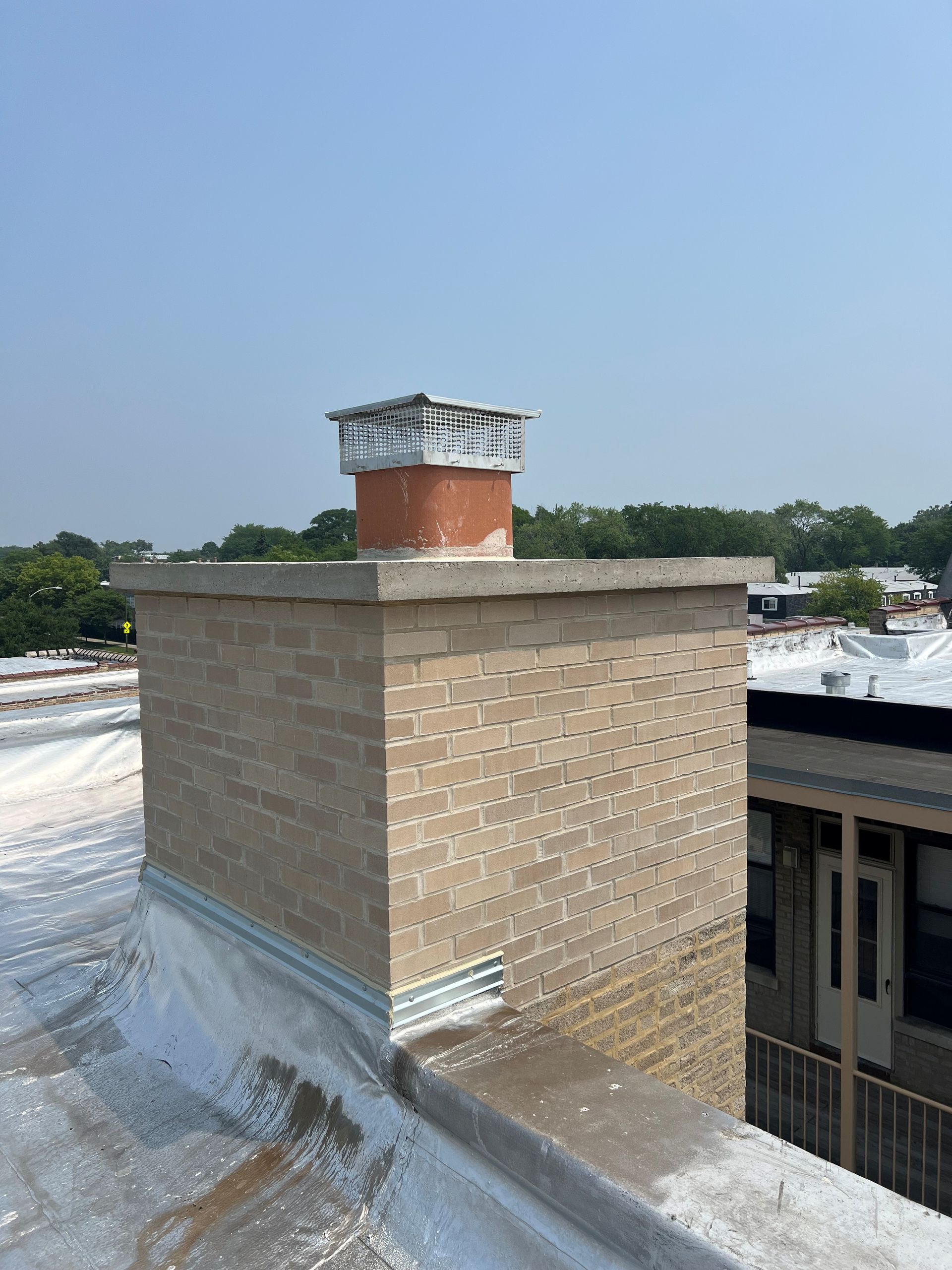 Brick rooftop chimney with metal cap on a flat roof under a clear blue sky.