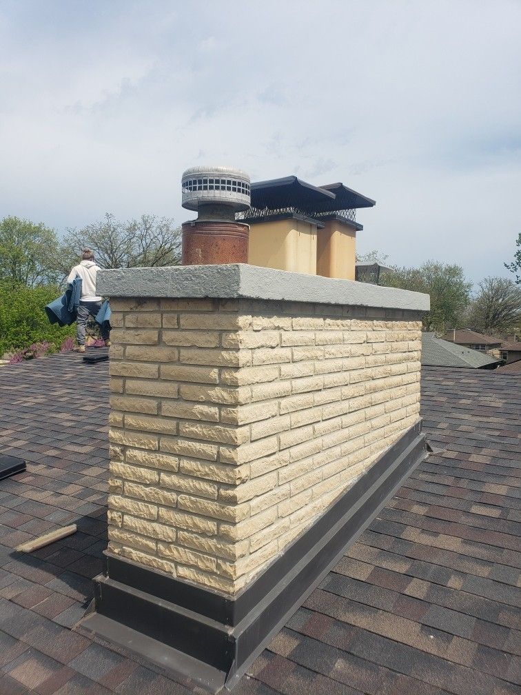 Rooftop chimney with cream brick sides, black base, and metal vent cap against a cloudy sky.