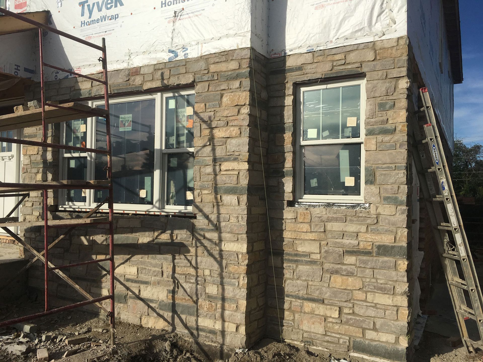 Stone house exterior under construction with scaffolding, windows, and Tyvek wrap at the roofline
