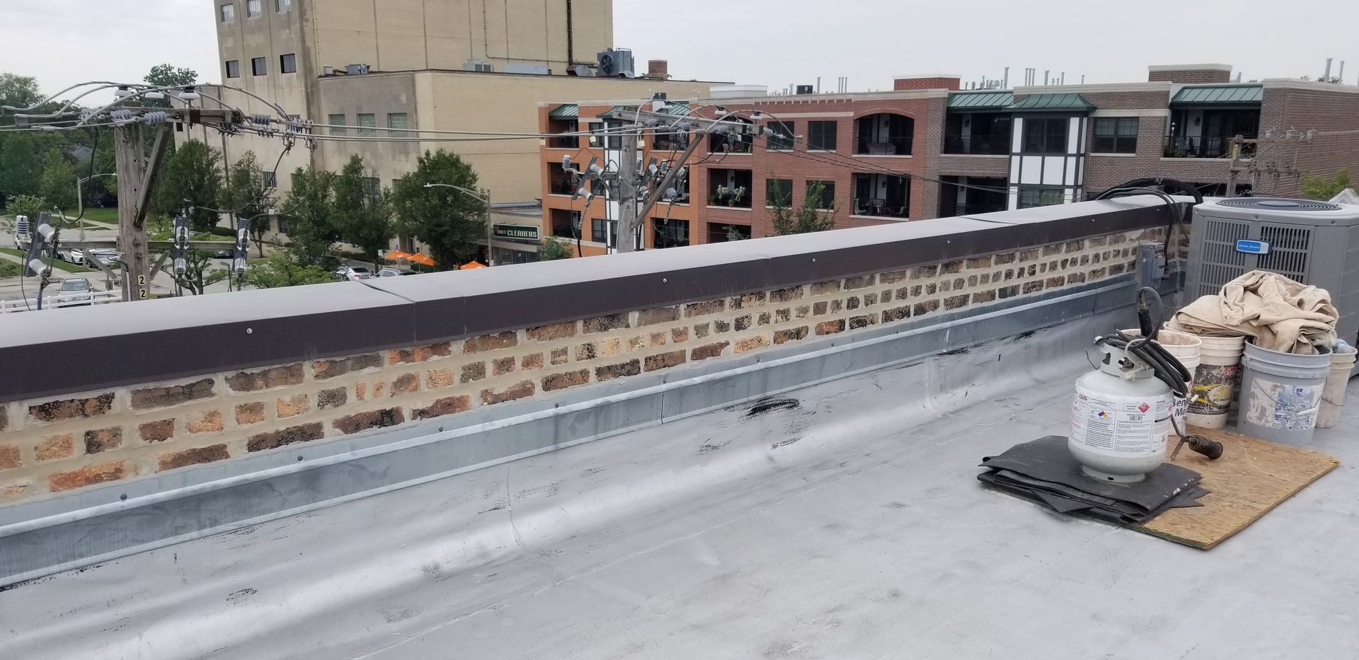 Rooftop with HVAC units and a low brick parapet, overlooking nearby city buildings.