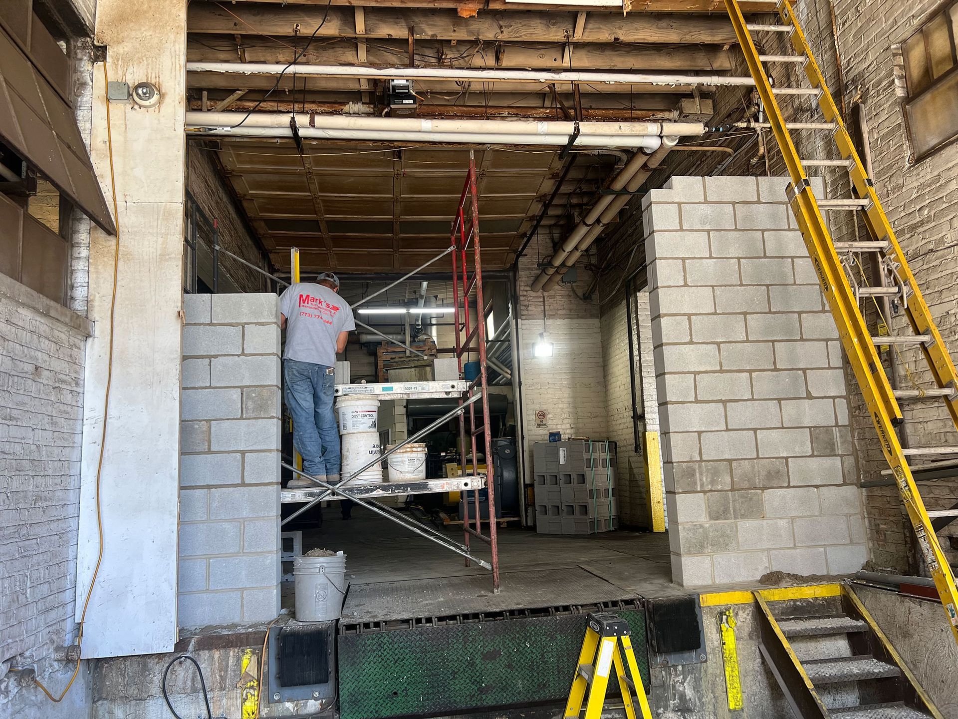 Construction site interior with workers on scaffolding, cinder block wall, and yellow ladder.