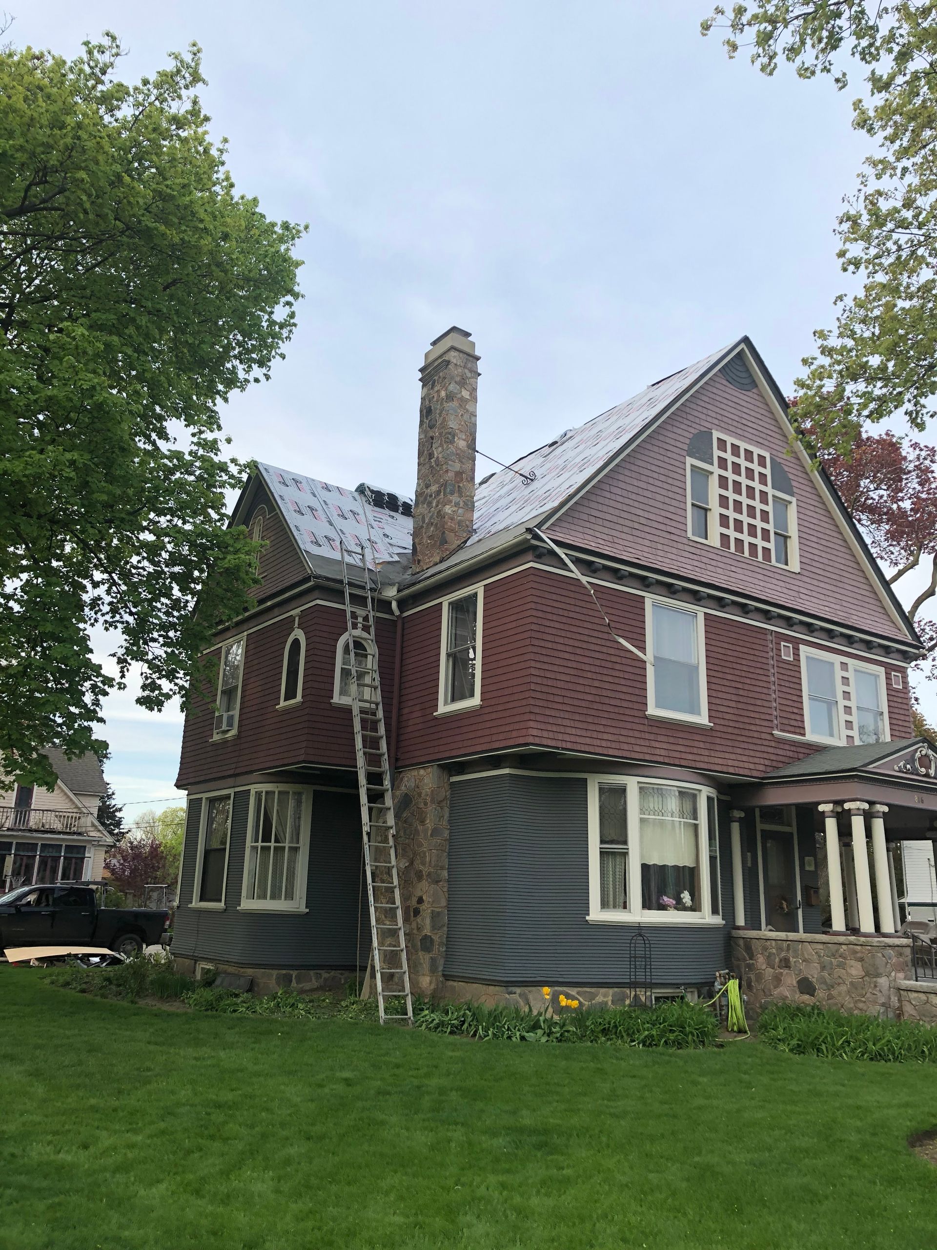 Damaged red-brick house with broken windows, ladder on chimney, and sagging siding in a grassy yard