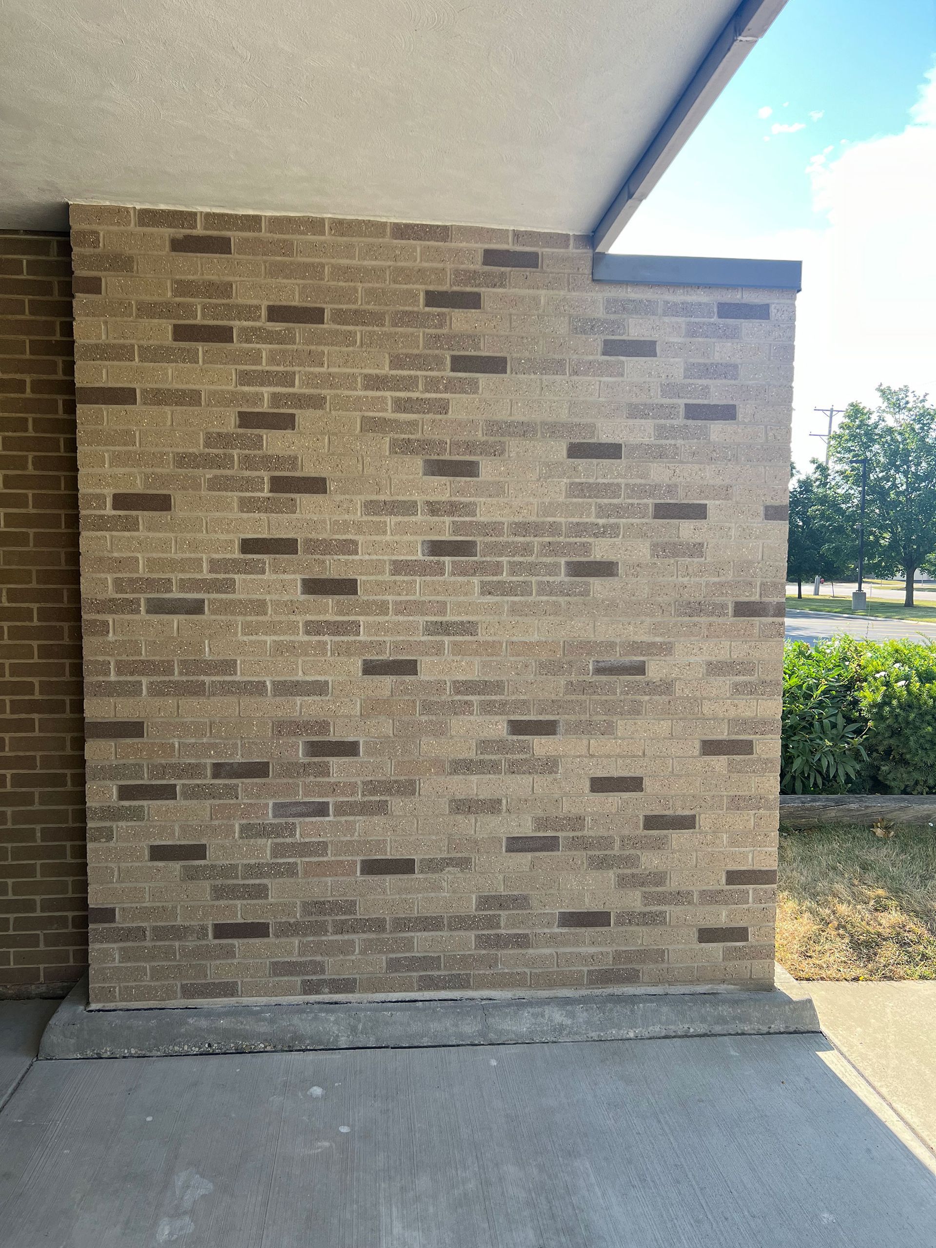 Brick wall beside a concrete walkway under a covered overhang, with shrubs and a tree in the background