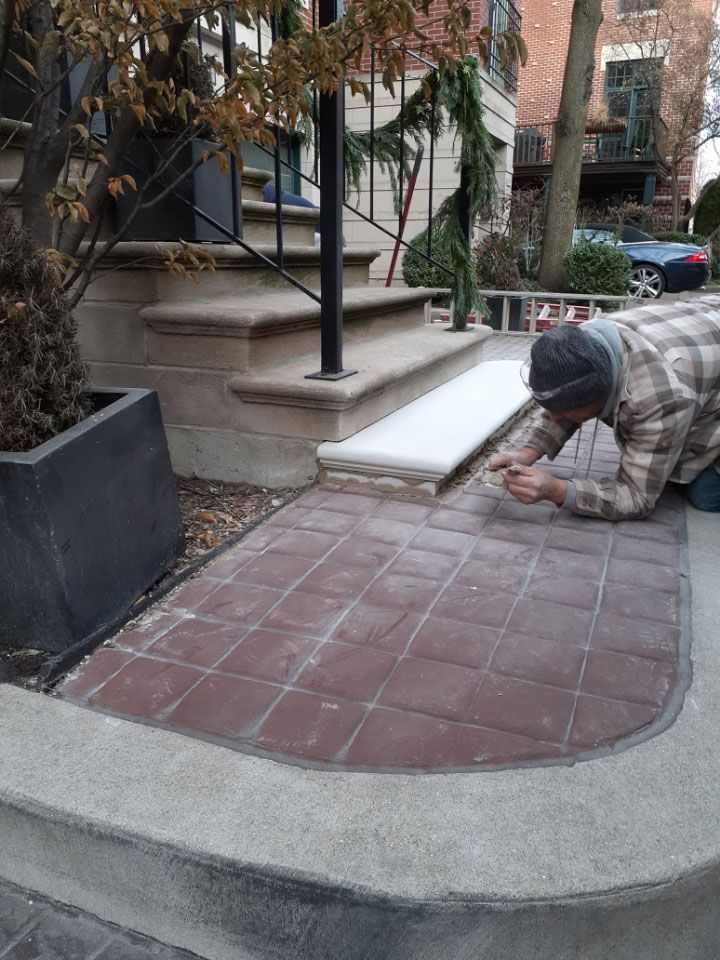 Worker kneeling to lay red pavers beside a curved concrete sidewalk in front of a building