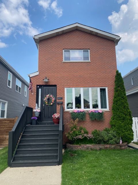 Two-story red brick house with black stairs, front lawn, and flower boxes under a blue sky