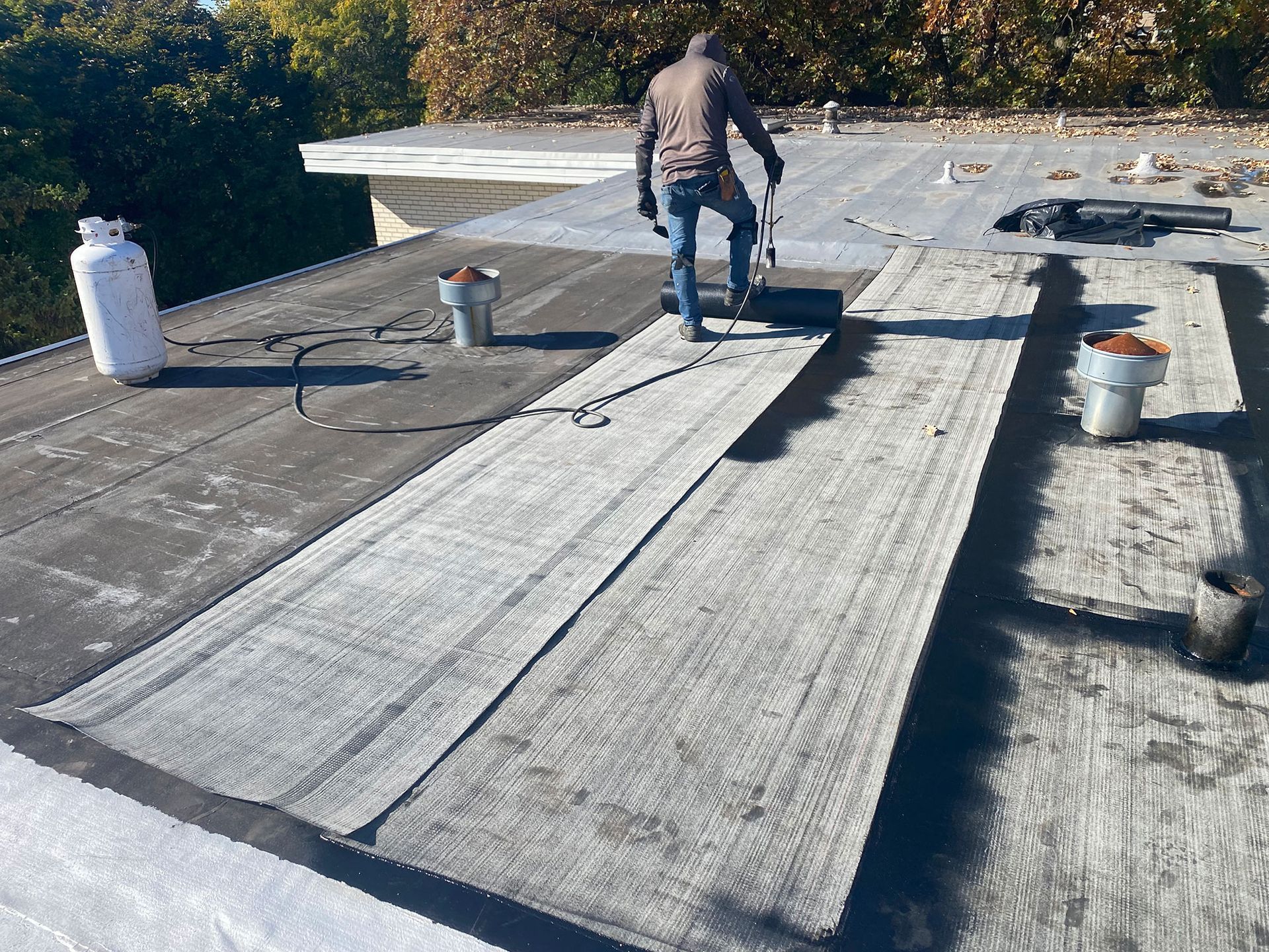 Worker installing roofing material on a flat roof, with rolled membrane and roof vents nearby.