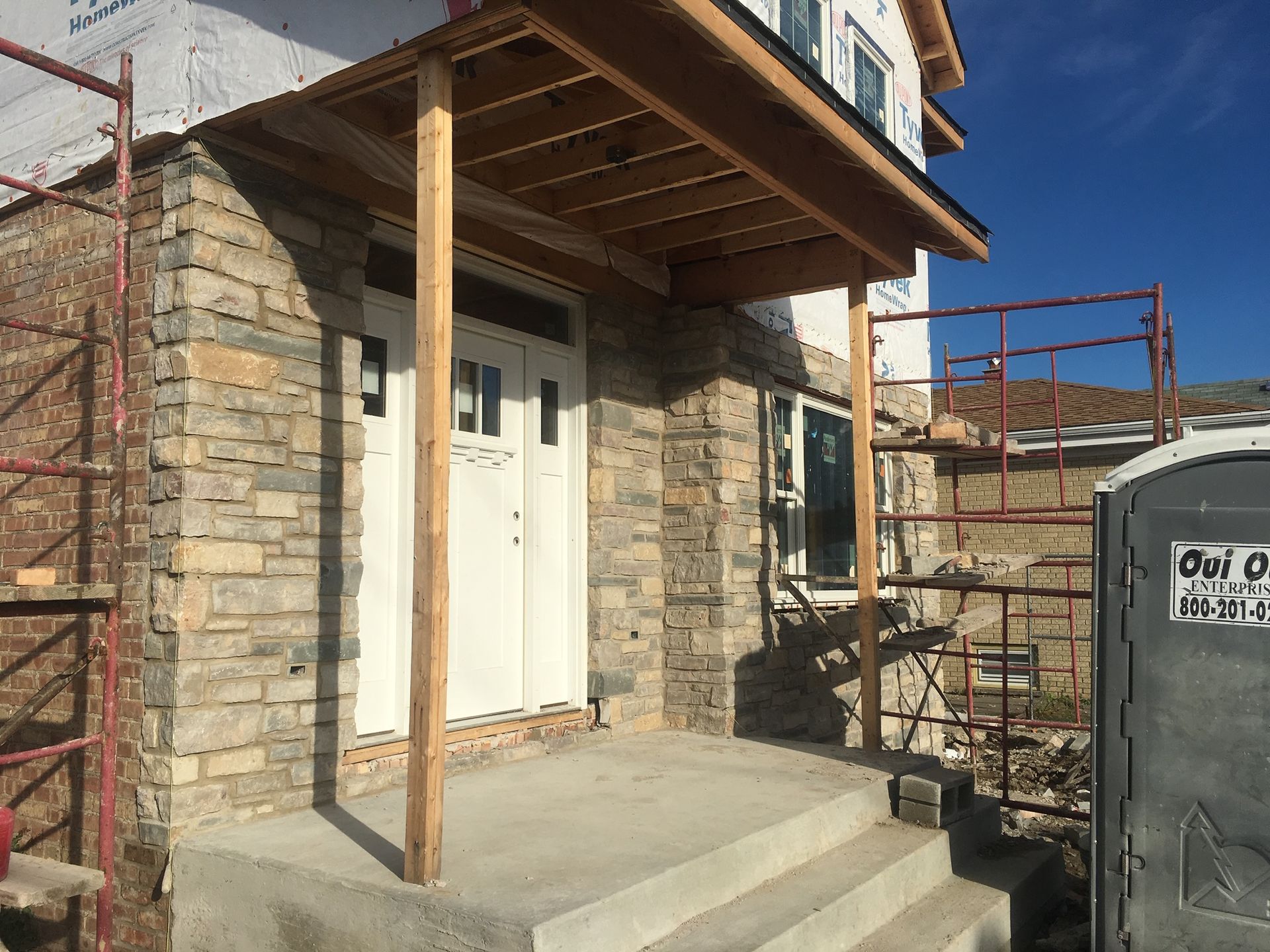 Stone house entrance with white door, small concrete porch, and wooden canopy under construction