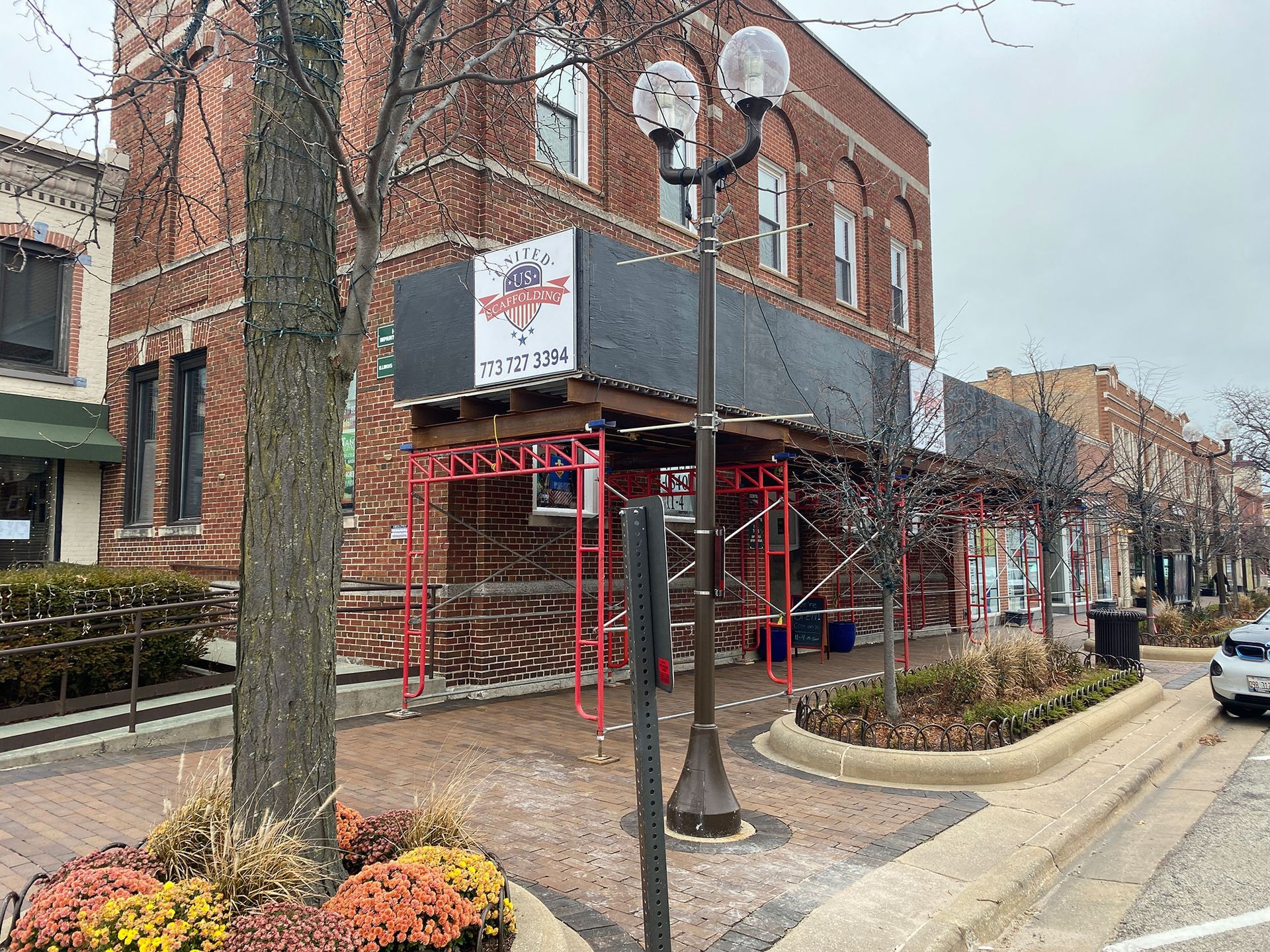 Scaffolded brick corner building on a city sidewalk with a hanging sign and bare trees along the street
