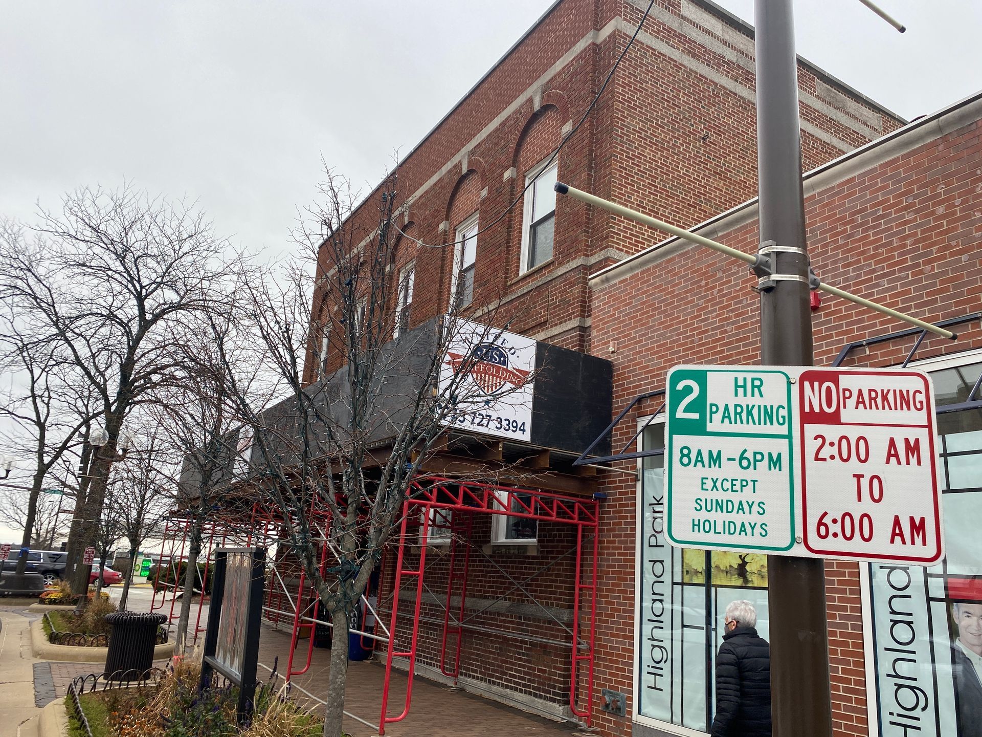Brick storefront on a city corner with a red awning, black gate, and several parking signs on the sidewalk