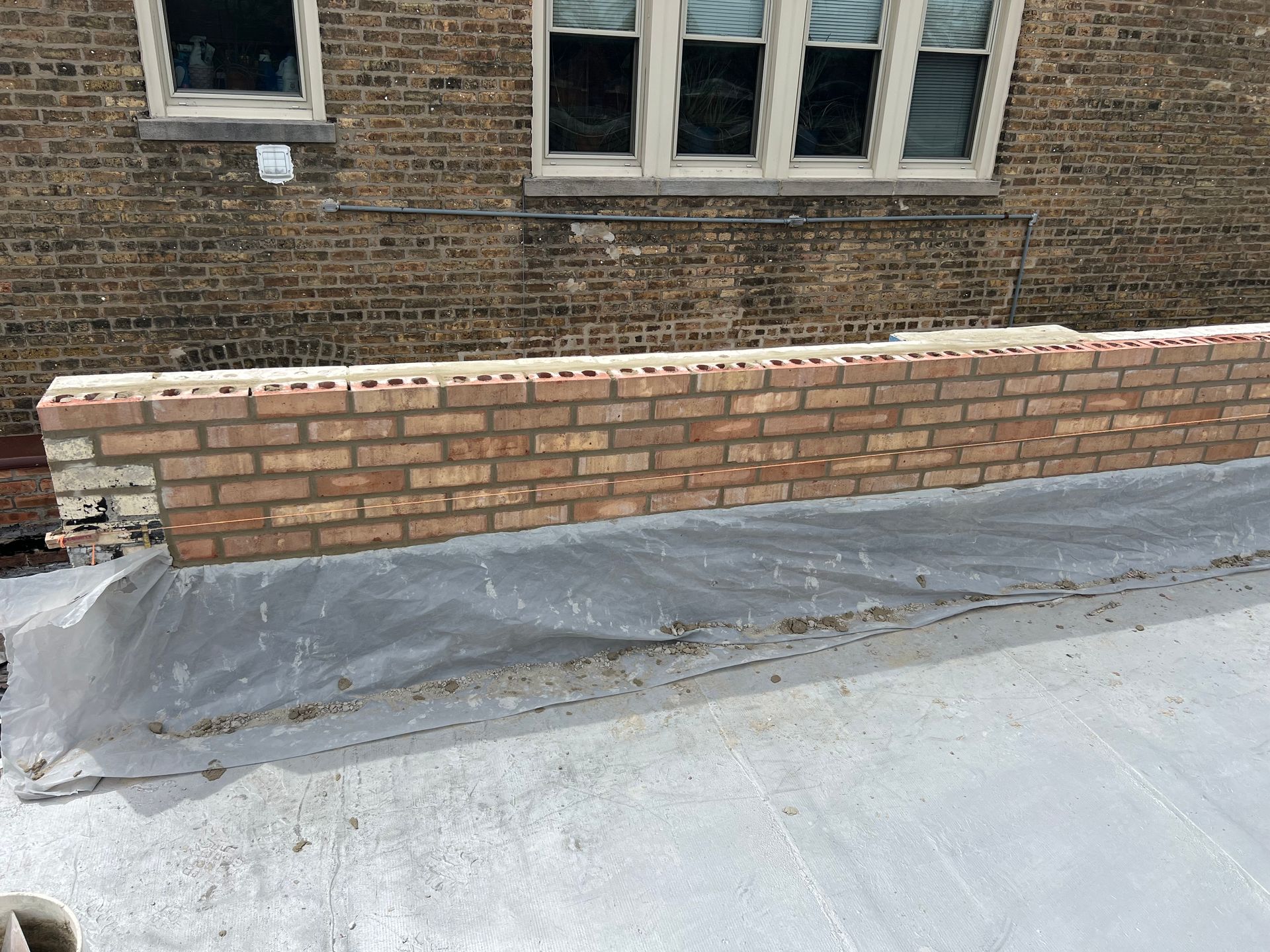 Brick wall under construction beside a brick building with gray tarp covering the ground