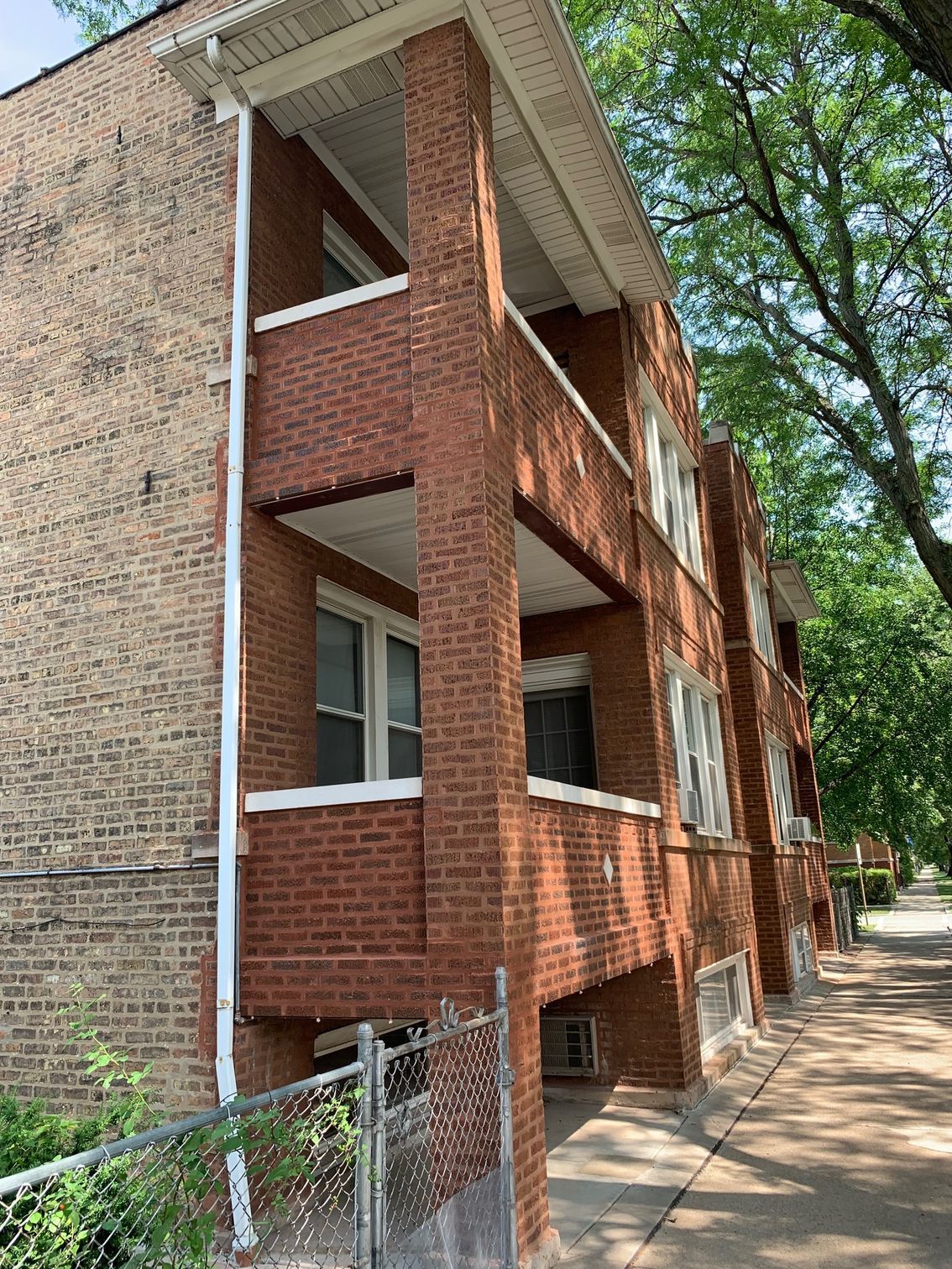 Brick apartment building with covered balconies beside a tree-lined sidewalk