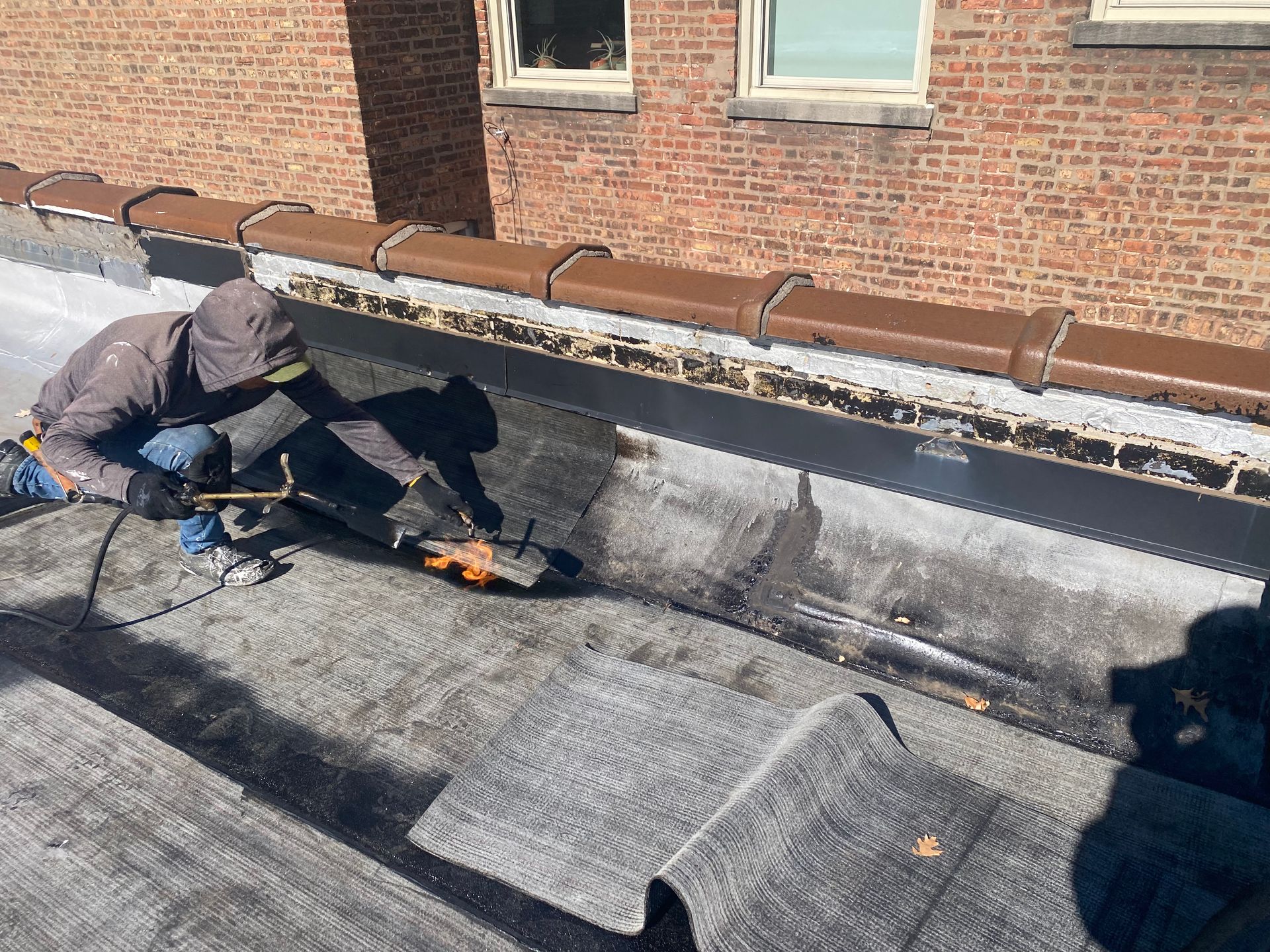 Worker repairing a flat roof beside a brick wall, using tools on gray roofing material.