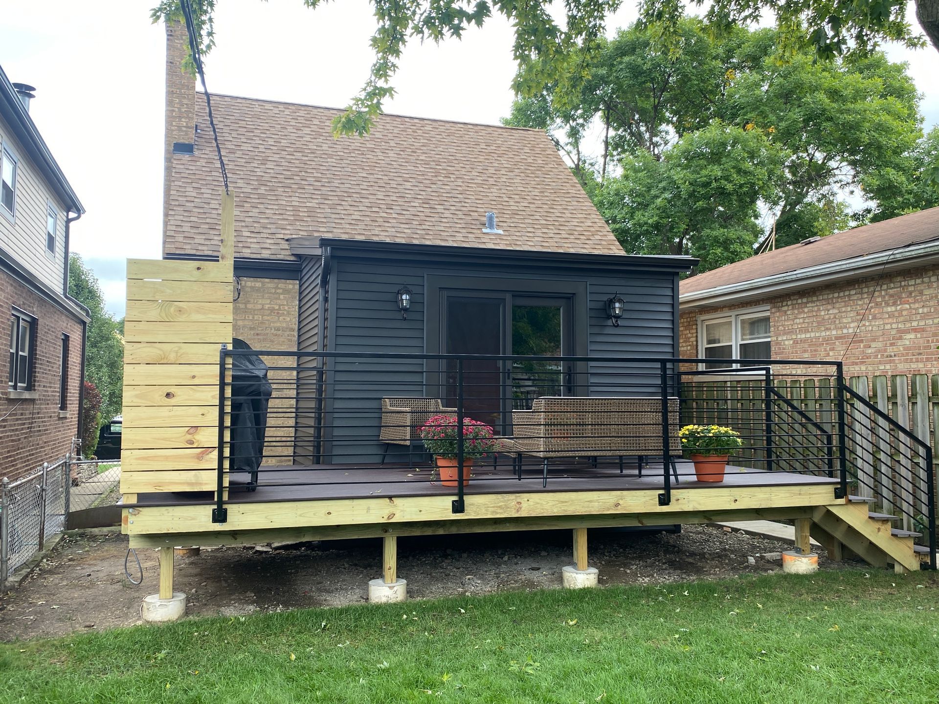Small dark gray backyard cabin with a wooden deck, black railing, and potted flowers in a grassy yard.