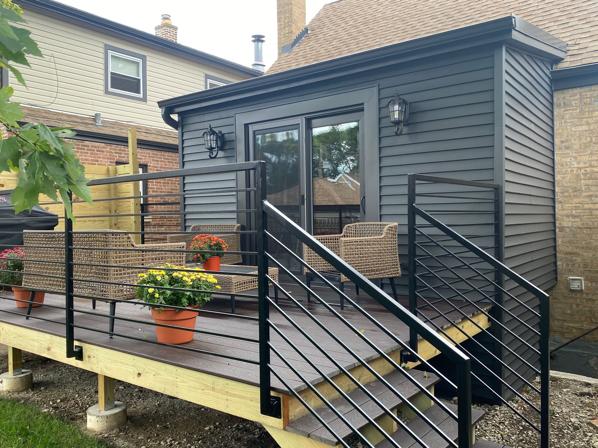 Small raised deck with black railings, stairs, and potted flowers beside a gray house.